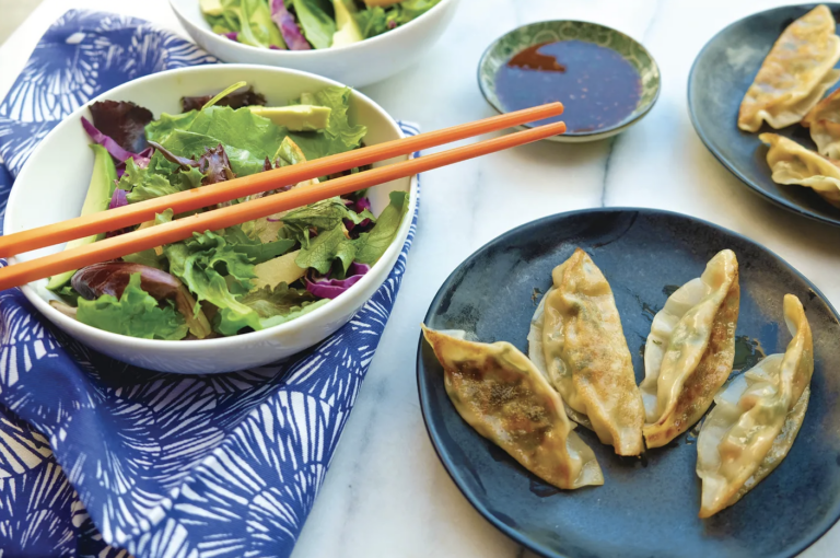 side salad of mixed lettuce in a white bowl with chopsticks balanced on top next to a blue plate with 4 potstickers on top